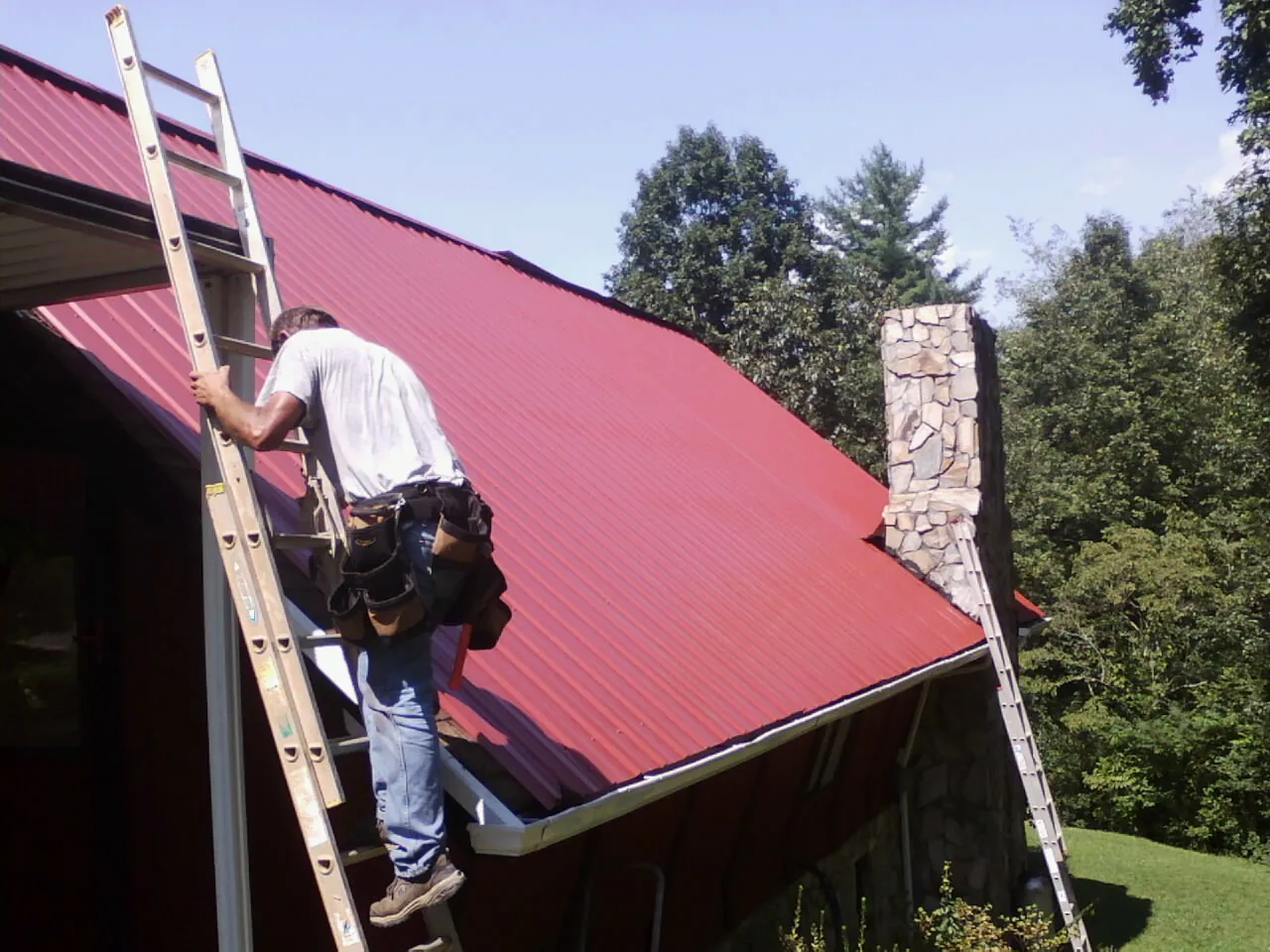 Roofer on a ladder beside a red metal roof and stone chimney — Soffit & Fascia Repair by Roofing Brothers in Norton